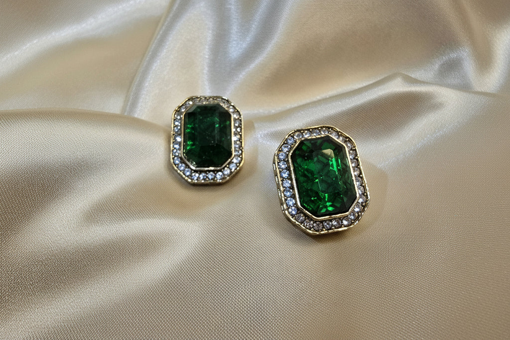 Pair of emerald green gemstone earrings with gold accents on a beige satin background